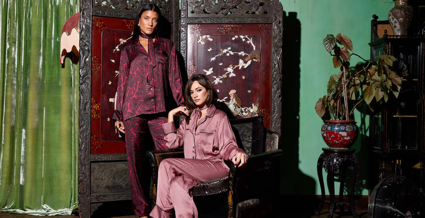 Two women in matching outfits sitting on a decorative chair in a room with green walls and various decor items.